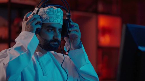 تحرير المشاهد وإضافة المؤثرات الصوتية والبصرية، تصوير مقرب لمونتير عربي خليجي عماني يرتدي الدشداشة و الكمة وسماعات الرأس يجلس داخل الاستديو يستخدم شاشة متعددة لتصميم الفيديوهات، مفهوم صناعة المحتوى الرقمي، ايماءات الاندماج و التركيز