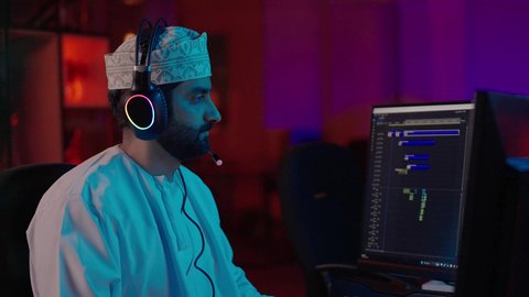 تحسين اللقطات وإدخال المؤثرات، تصوير مقرب من الجانب لمونتير عربي خليجي عماني يرتدي الدشداشة و الكمة وسماعات الرأس يجلس داخل الاستديو يستخدم شاشة متعددة لتصميم الفيديوهات، مفهوم صناعة المحتوى الرقمي، ايماءات الاندماج و التركيز