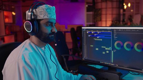 تعديل المقاطع وإضافة اللمسات الإبداعية، تصوير مقرب من الجانب لمونتير عربي خليجي عماني يرتدي الدشداشة و الكمة وسماعات الرأس يجلس داخل الاستديو يستخدم شاشة متعددة لتصميم الفيديوهات، مفهوم صناعة المحتوى الرقمي، ايماءات الاندماج و التركيز