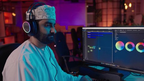 تعديل الفيديوهات وإضافة التأثيرات البصرية، تصوير مقرب من الجانب لمونتير عربي خليجي عماني يرتدي الدشداشة و الكمة وسماعات الرأس يجلس داخل الاستديو يستخدم شاشة متعددة لتصميم الفيديوهات، مفهوم صناعة المحتوى الرقمي، النقر على لوحه المفاتيح