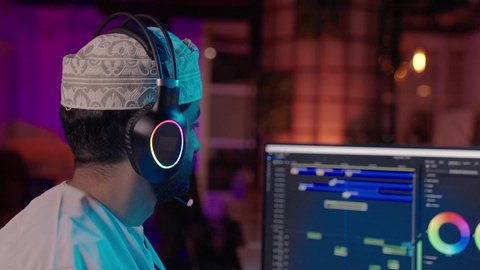 تحرير اللقطات وإدراج المؤثرات، تصوير مقرب من الجانب لمونتير عربي خليجي عماني يرتدي الدشداشة و الكمة وسماعات الرأس يجلس داخل الاستديو يستخدم شاشة متعددة لتصميم الفيديوهات، مفهوم صناعة المحتوى الرقمي