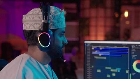 تعديل المشاهد وإضافة المؤثرات، تصويرمقرب من الجانب لمونتير عربي خليجي عماني يرتدي الدشداشة و الكمة وسماعات الرأس يجلس داخل الاستديو يستخدم شاشة متعددة لتصميم الفيديوهات، مفهوم صناعة المحتوى الرقمي