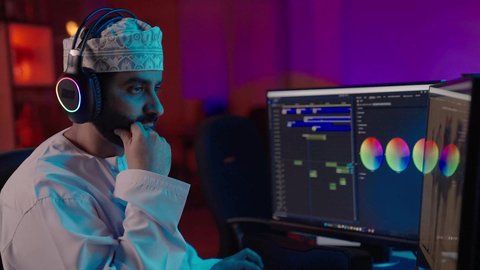 مفهوم الإنتاج التلفزيوني والسينمائي، تصوير من الجانب لمونتير عربي خليجي عماني يرتدي الدشداشة و الكمة وسماعات الرأس يجلس داخل الاستديو يعمل على ترتيب المشاهد باستخدام برامج الحاسوب المتطورة يضع يدية على ذقنه بايماءات التفكير و التركيز