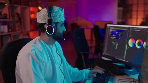 صناعة المحتوى الإعلامي، تصوير من الجانب لمونتير عربي خليجي عماني يرتدي الدشداشة و الكمة وسماعات الرأس يجلس داخل الاستديو يعمل على ترتيب المشاهد باستخدام برامج الحاسوب المتطورة، ايماءات الاندماج و التركيز