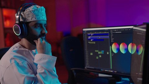 مونتاج وإخراج مرئي، تصوير من الجانب لمونتير عربي خليجي عماني يرتدي الدشداشة و الكمة وسماعات الرأس يجلس داخل الاستديو يعمل على ترتيب المشاهد باستخدام برامج الحاسوب المتطورة، ايماءات الاندماج و التركيز