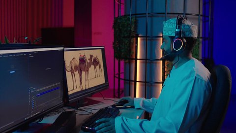 الاستعداد لعرض الأعمال الفنية، ايماءات الاندماج و التركيز، تصوير من الجانب لمونتير عربي خليجي عماني يرتدي الدشداشة و الكمة وسماعات الرأس يجلس داخل الاستديو يركز على شاشة المونتاج، استخدام برامج تحرير متطورة لإنتاج الفيديوهات، النقر على لوحه المفاتيح