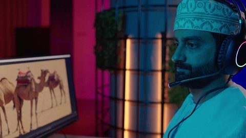 صناعة الأفلام والبرامج،ايماءات الاندماج و التركيز، تصويرمقرب من الجانب لمونتير عربي خليجي عماني يرتدي الدشداشة و الكمة وسماعات الرأس يجلس داخل الاستديو يركز على شاشة المونتاج، استخدام برامج تحرير متطورة لإنتاج الفيديوهات