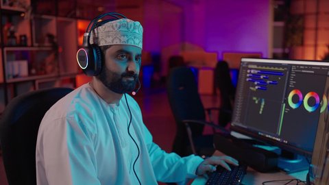 ايماءات الاندماج و التركيز ، مفهوم صناعة الأفلام والإخراج، تصوير من الجانب لمونتير عربي خليجي عماني يرتدي الدشداشة و الكمة وسماعات الرأس يجلس داخل الاستديو يركز على شاشة المونتاج، استخدام برامج تحرير متطورة لإنتاج الفيديوهات