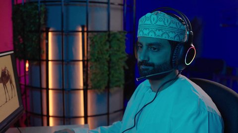 مفهوم صناعة الأفلام والإخراج، تصويرمقرب من الجانب لمونتير عربي خليجي عماني يرتدي الدشداشة و الكمة وسماعات الرأس يجلس داخل الاستديو يركز على شاشة المونتاج، استخدام برامج تحرير متطورة لإنتاج الفيديوهات