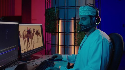 الاستفادة من برامج وتقنيات المونتاج الحديثة، تصوير من الجانب لمونتير عربي خليجي عماني يرتدي الدشداشة و الكمة وسماعات الرأس يجلس داخل الاستديو يقوم بتعديل الصوت على شاشة الكمبيوتر، صناعة الأفلام والبرامج التلفزيونية