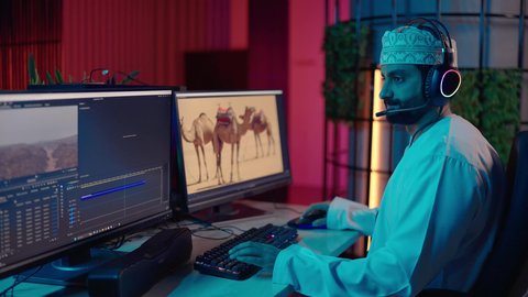 مفهوم صناعة المحتوى المرئي، النقر على لوحه المفاتيح، تصوير من الجانب لمونتير عربي خليجي عماني يرتدي الدشداشة و الكمة وسماعات الرأس يجلس داخل الاستديو يتابع شاشة المونتاج لمراجعة مقاطع الفيديو وإنتاج الأفلام، دمج المشاهد والمؤثرات المرئية