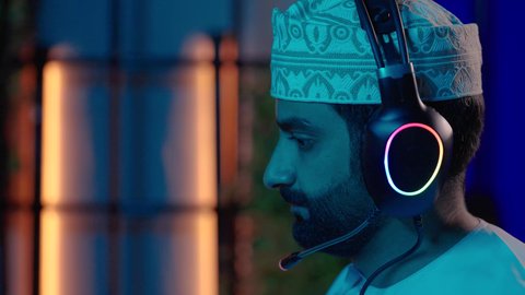 المونتاج والإبداع الفني، تصوير من الجانب لمونتير عربي خليجي عماني يرتدي الدشداشة و الكمة وسماعات الرأس يجلس داخل الاستديو يركز على شاشة الكمبيوتر لإخراج مشهد متكامل، التحكم في الانتقالات والمؤثرات