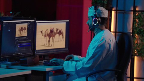 تعديل المشاهد وإضافة المؤثرات، تصويرمن الجانب لمونتيرعربي خليجي عماني يرتدي الدشداشة و الكمة وسماعات الرأس يجلس داخل الاستديو يستخدم شاشة متعددة لتصميم الفيديوهات، مفهوم صناعة المحتوى الرقمي، النقر على لوحه المفاتيح