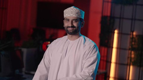 مفهوم الأناقة والرجولة، الاهتمام بالمظهر الخارجي، تصوير لمونتيرعربي خليجي عماني يرتدي الدشداشة و الكمة داخل الاستديو يقف باستقامة بأيدي متقاطعة ، النظر الى الكاميرا بايماءات السعادة و السرور