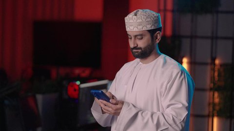 التواصل مع الأصدقاء، تصوير لمونتير عربي خليجي عماني يرتدي الدشداشة و الكمة داخل الاستديو يستخدم الهاتف المحمول، تصفح الإنترنت عبر الجوال ، إستخدام وسائل التواصل الإجتماعي، استخدام تقنيات حديثة