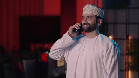 ايماءات السعادة و السرور، متابعة الأعمال والتواصل أثناء إنتاج الفيديو، تصوير لمونتير عربي خليجي عماني يرتدي الدشداشة و الكمة يعمل في مجال الميديا داخل غرفة المونتاج يستخدم الهاتف المحمول