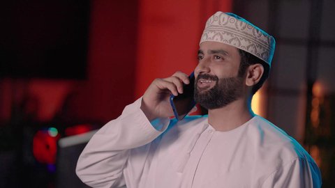 متابعة الأعمال والتواصل أثناء إنتاج الفيديو، تصويرمقرب لمونتير عربي خليجي عماني يرتدي الدشداشة و الكمة يعمل في مجال الميديا داخل غرفة المونتاج يستخدم الهاتف المحمول، ايماءات السعادة و السرور