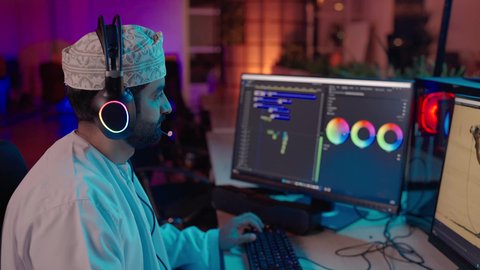 تعديل المشاهد وإضافة المؤثرات، تصوير من الجانب لمونتير عربي خليجي عماني يرتدي الدشداشة و الكمة و سماعات الرأس يستخدم شاشة متعددة لتصميم الفيديوهات، مفهوم صناعة المحتوى الرقمي ، النقر على لوحه المفاتيح