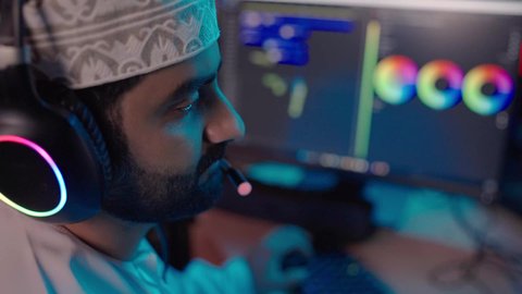 بيئة استوديو حديثة، صناعة الأفلام والبرامج، تصوير مقرب من الجانب لمونتير عربي خليجي عماني يرتدي الدشداشة و الكمة و سماعات الرأس يتابع الصوت والصورة على شاشة الكمبيوتر