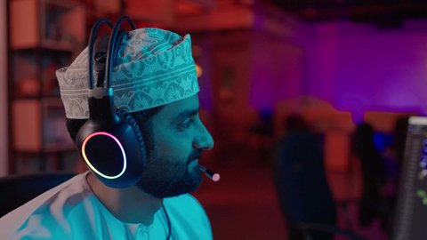 صناعة الأفلام والبرامج، تصوير مقرب من الجانب لمونتير عربي خليجي عماني يرتدي الدشداشة و الكمة و سماعات الرأس يتابع الصوت والصورة على شاشة الكمبيوتر، بيئة استوديو حديثة