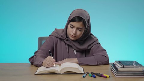 مراجعة الدروس اليومية، ثقافة التعلم الذاتي، قضاء الوقت بالتعلم والتثقف، الاستمتاع بقراءة الكتب والقصص والروايات، بورتريه لشابة عربية خليجية سعودية محجبة ترتدي العباءة تجلس خلف مقعد الدراسة تقوم بالمذاكرة والاجتهاد والدراسة، خلفية زرقاء