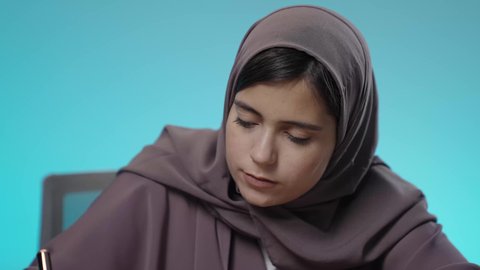 جودة التعليم في المملكة، مراجعة الدروس اليومية، ثقافة التعلم الذاتي، قضاء الوقت بالتعلم والتثقف، الاستمتاع بقراءة الكتب والقصص والروايات، بورتريه مقرب لشابة عربية خليجية سعودية محجبة ترتدي العباءة تجلس خلف مقعد الدراسة تقوم بالمذاكرة والاجتهاد والدراسة، خلفية زرقاء