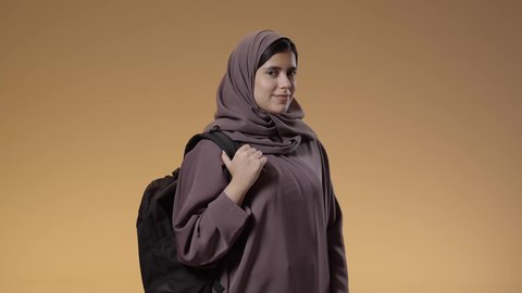 Looking at the camera with confident gestures, portrait of a Saudi Arabian Gulf university student carrying her university bag, preparing for the new university year, a diligent student eager to go to university, education in schools and universities in the Kingdom of Saudi Arabia, blue background.