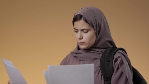 Education in schools and universities in the Kingdom, gestures of discontent and anger, eagerness to go to university, preparation for the new academic year, close-up portrait of a Saudi Arabian Gulf female university student carrying a university bag and holding white papers in her hands, studying, hard work and studying, blue background.