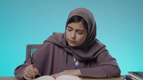 The culture of self-learning, studying, hard work and studying, spending time learning and becoming educated, enjoying reading books, stories and novels, a portrait of a veiled Saudi Arabian Gulf young woman wearing an abaya sitting behind a study desk reading a book, brown background