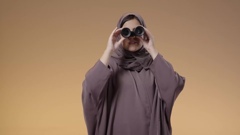 الرؤية المستقبلية وتحديد الأهداف والطموحات، المراقبة ومشاهدة الأشياء البعيدة، بورتريه لامرأة عربية خليجية سعودية محجبة ترتدي العباءة التقليدية تمسك بيديها منظار ثنائي العينين وتنظر من خلاله، خلفية بني