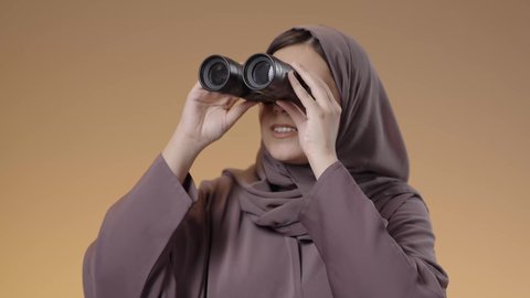 Observation and watching distant things, future vision and setting goals and ambitions, close-up portrait of a veiled Saudi Arabian Gulf woman wearing an abaya holding binoculars in her hands and looking through them, brown background