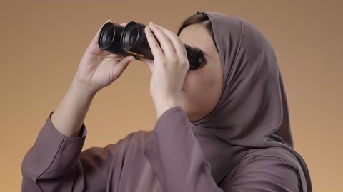 Observation and watching distant things, future vision and setting goals and ambitions, close-up portrait of a veiled Saudi Arabian Gulf woman wearing an abaya holding binoculars in her hands and looking through them, brown background