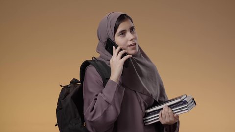 استخدام جهاز تقني حديث ومتطور، بورتريه لطالبة عربية خليجية سعودية محجبة ترتدي العباءة وحقيبة الظهر تحمل الكتب الجامعية، إجراء مكالمات عبر الهاتف المحمول، التواصل والدردشة مع الأصدقاء، خلفية بني