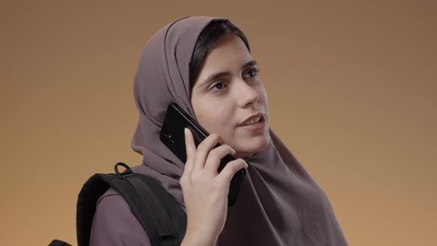 إجراء مكالمات عبر الهاتف المحمول، استخدام جهاز تقني حديث ومتطور، بورتريه مقرب لطالبة عربية خليجية سعودية محجبة ترتدي العباءة وحقيبة الظهر الجامعية، التواصل والدردشة مع الأصدقاء، خلفية بني