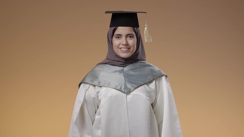 أهمية الدراسة الجامعية، التعليم في جامعات المملكة العربية السعودية، النجاح وتحقيق الاهداف، اللباس الاكاديمي المتميز، بورتريه لطالبة عربية خليجية سعودية ترتدي لباس التخرج تحمل بيدها تنظر الى الكاميرا بإيماءات الفرح والسرور، خلفية بني