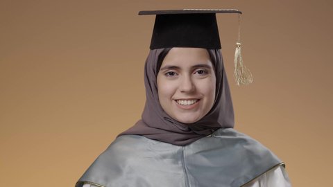 مفهوم النجاح وتحقيق الاهداف، اللباس الاكاديمي المتميز، أهمية الدراسة الجامعية، التعليم في جامعات المملكة العربية السعودية، بورتريه مقرب لطالبة عربية خليجية سعودية ترتدي لباس التخرج تحمل بيدها تنظر الى الكاميرا بإيماءات الفرح والسرور، خلفية بني