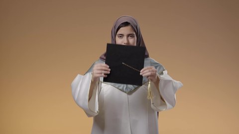 وضع قبضة التخرج، جودة التعليم في جامعات المملكة، مفهوم النجاح وتحقيق الاهداف، اللباس الاكاديمي المتميز، أهمية الدراسة الجامعية، بورتريه لطالبة عربية خليجية سعودية ترتدي لباس التخرج تحمل بيدها تنظر الى الكاميرا بإيماءات الفرح والسرور، خلفية بني