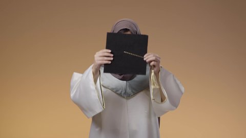 الوقوف بأيدي متقاطعة، وضع قبضة التخرج، التعليم في جامعات المملكة، مفهوم النجاح وتحقيق الاهداف، اللباس الاكاديمي المتميز، أهمية الدراسة الجامعية، بورتريه لطالبة عربية خليجية سعودية ترتدي لباس التخرج تحمل بيدها تنظر الى الكاميرا بإيماءات الفرح والسرور، خلفية بني