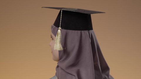 جودة التعليم في جامعات المملكة العربية السعودية، مفهوم النجاح وتحقيق الاهداف، اللباس الاكاديمي المتميز، أهمية الدراسة الجامعية، بورتريه مقرب لطالبة عربية خليجية سعودية ترتدي لباس التخرج تحمل بيدها تنظر الى الكاميرا بإيماءات الفرح والسرور، خلفية بني