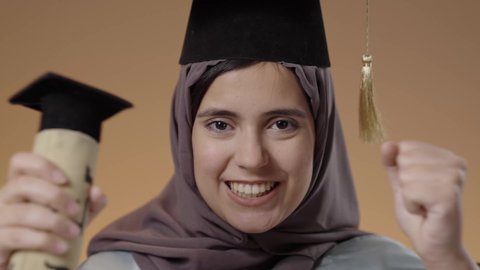 رفع قبضة اليدين بإيماءات النجاح وتحقيق الاهداف، اللباس الاكاديمي المتميز، بورتريه مقرب لطالبة عربية خليجية سعودية ترتدي لباس التخرج تحمل بيدها الشهادة تنظر الى الكاميرا بإيماءات الفرح والسرور، التعليم في جامعات المملكة العربية السعودية، خلفية بني