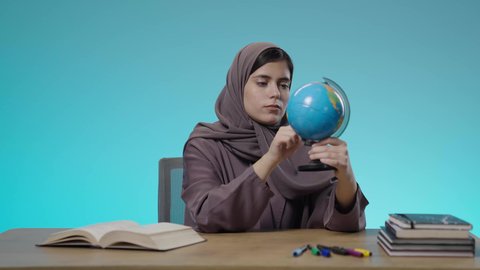 دراسة العلوم الجغرافية، توسيع الآفاق وفهم التنوع الثقافي والطبيعي، الاهتمام في التعليم لتطوير المجتمع السعودي، طالبة عربية خليجية سعودية ترتدي الزي التقليدي وتقوم بتدوير مجسم الكرة الأرضية بإصبعها ايماءات التركيز والتمحيص، خلفية زرقاء.