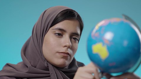 ر توسيع الآفاق وفهم التنوع الثقافي والطبيعي، دراسة العلوم الجغرافية، الاهتمام في التعليم لتطوير المجتمع السعودي، بورتريه مقرب لطالبة عربية خليجية سعودية ترتدي الزي التقليدي وتقوم بتدوير مجسم الكرة الأرضية بإصبعها ايماءات التركيز والتمحيص، خلفية زرقاء.