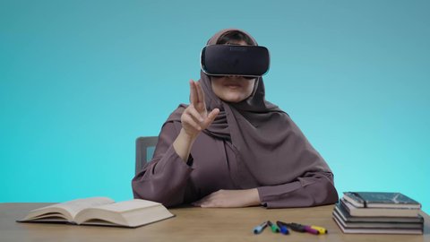 نشاط دراسي باستعمال الواقع الافتراضي، بورتريه لطالبة جامعية عربية خليجية سعودية تجلس على الكرسي ترتدي نظارات (في آر) ثلاثية الأبعاد ، جودة التعليم المتطور في السعودية، الدراسة عبر منصة تعليمية للتعلم عن بعد، استخدام حاسة اللمس لتحريك العناصر الهولوجرامية، خلفية زرقاء