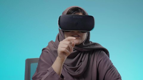 استخدام حاسة اللمس لتحريك العناصر الهولوجرامية ، بورتريه لشابة عربية خليجية سعودية محجبة تجلس على الكرسي ترتدي نظارات الواقع الإفتراضي (في آر) ثلاثية الأبعاد ، المتعة والتسليه والترفيه، مفهوم التقنيات الإلكترونية الحديثة، خلفية زرقاء