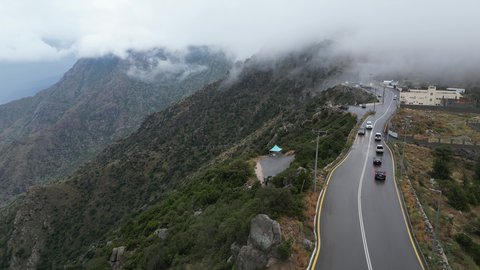 The rocky mountain ranges of Abha and Namas, the rough terrain and stone roads, aerial photography of the mountainous environment in the Asir region, the white clouds and foggy atmosphere covering the high peaks, the green nature and beautiful parks and gardens, the trees and plants on the mountain slopes