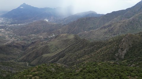 The rocky mountain ranges of Abha and Namas, the rough terrain and stone roads, aerial photography of the mountainous environment in the Asir region, the white clouds and foggy atmosphere covering the high peaks, the green nature and beautiful parks and gardens, the trees and plants on the mountain slopes