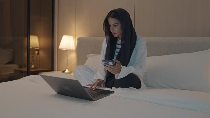 الفيام بمكالمة فيديو, ايماءات تدل على التحية والسلام, شابة عربية خلجية سعودية تجلس في غرفة النوم على السرير  تقوم باستخدام الحاسوب المحمول, الدردشة و التواصل الاجتماعي, تصفح الانترنت واستخدام تقنيات حديثة, اجراء بث مباشر
