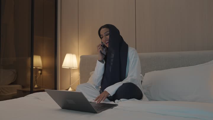 الفيام بمكالمة فيديو, ايماءات تدل على التحية والسلام, شابة عربية خلجية سعودية تجلس في غرفة النوم على السرير تقوم باستخدام الحاسوب المحمول, الدردشة و التواصل الاجتماعي, تصفح الانترنت واستخدام تقنيات حديثة, اجراء بث مباشر