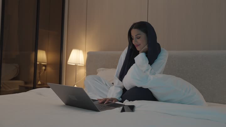 Making a video call, gestures indicating greeting and peace, a young Saudi Arabian Gulf woman sitting in the bedroom on the bed using the laptop, chatting and socializing, surfing the internet and using modern technologies, conducting a live broadcast