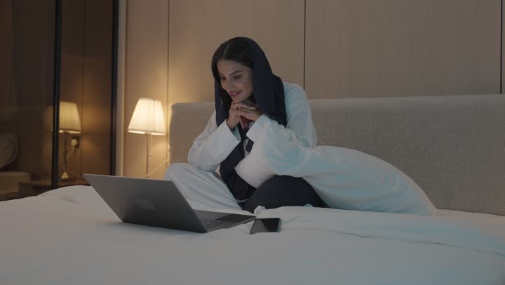 Making a video call, gestures indicating greeting and peace, a young Saudi Arabian Gulf woman sitting in the bedroom on the bed using the laptop, chatting and socializing, surfing the internet and using modern technologies, conducting a live broadcast